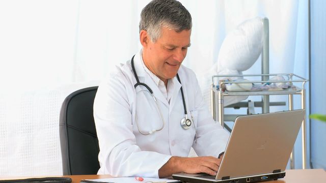 Doctor wearing white coat and stethoscope, sitting at desk and typing on laptop. Modern medical office with medical equipment in the background. Ideal for content related to healthcare technology, medical consulting, digital health records, and telemedicine services.