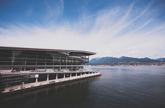 The image depicts a modern building by the waterfront, offering a striking contrast with majestic mountains in the background. The scene is peaceful with a clear sky and deep, tranquil water reflecting both the architecture and natural beauty. Ideal for use in urban planning presentations, travel brochures, and websites promoting tourism or real estate with a focus on serene environments and impressive views.