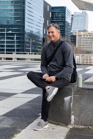 Middle-aged professional resting on urban ledge using phone with backpack and bottle