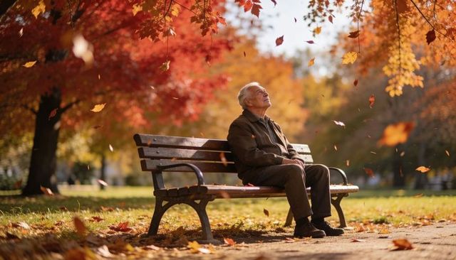 Senior man sitting on park bench amid falling autumn leaves, gazing upward in sunlight