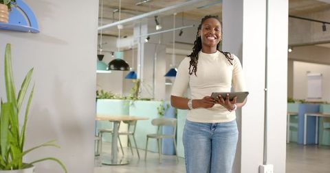 Smiling african woman holding tablet in coworking lounge wearing sweater and jeans