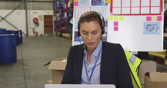 Woman warehouse supervisor wearing a headset and safety vest, working intently on a laptop. Ideal for use in articles, blogs, or training materials about logistics, productivity, warehouse management, and industrial work environments.