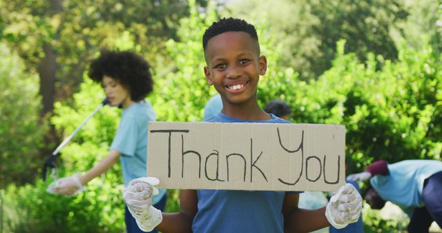 Smiling african american son holding thank you sign, clearing up trash outdoors with family. Ecology, volunteering, recycling, nature conservation, family and togetherness.