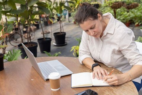 Mature woman writing in notebook at greenhouse table, laptop and coffee, nursery workspace