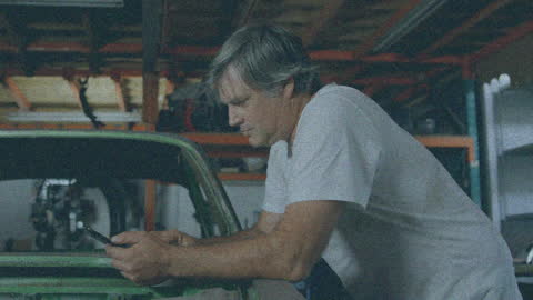 Older Man in Garage Using Smartphone Beside Classic Car