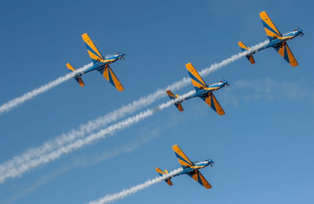 Several aerobatic planes flying in a close formation against a clear blue sky. The planes are leaving white smoke trails, demonstrating their synchronized flight skills and precision. This image is ideal for illustrating teamwork, aviation artistry, airshows, and pilots' skills. It is suitable for articles, advertisements, and other media related to aviation events, flying clubs, and aeronautics education.