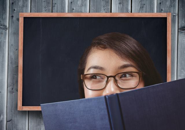 Girl in spectacles holding book on her mouth with chalkboard in background at classroom
