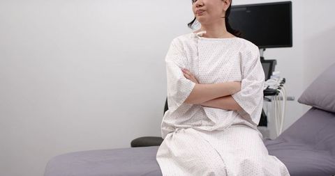 Woman sitting on exam table crossing arms wearing hospital gown in clinic with ultrasound
