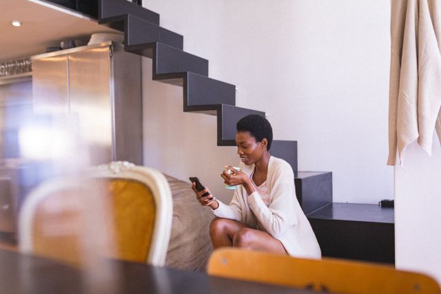 African american young woman drinking coffee while using smartphone in living room at home. unaltered, people, drink, lifestyle, home and technology concept.