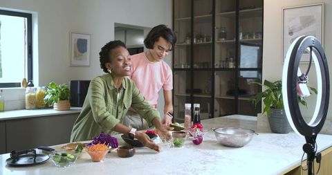 Diverse friends filming cooking tutorial at kitchen island, preparing fresh vegetable salad