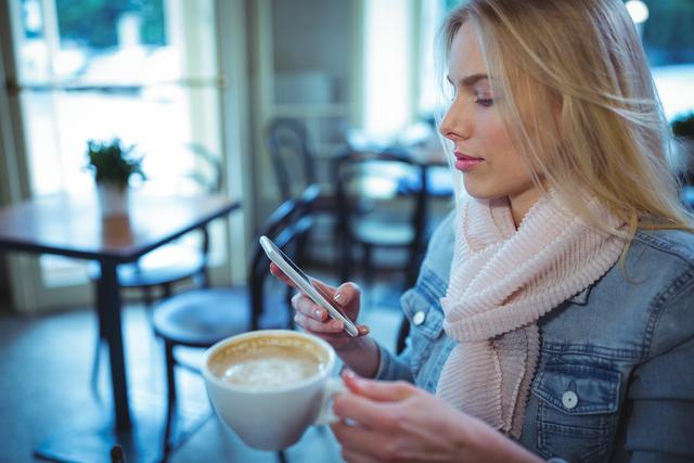 Blonde woman using a mobile phone while enjoying a large cup of coffee in a cozy café. She is wearing a denim jacket and a winter scarf, creating a casual and relaxed atmosphere. This scene is ideal for illustrating themes of modern lifestyle, technology use, social media engagement, urban living, and leisure activities. Perfect for use in advertisements, blog posts, social media content, and lifestyle articles.