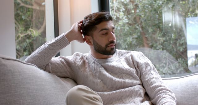 A man wearing a comfortable sweater is sitting on a couch, appearing lost in thought. Surrounded by the tranquility of a cozy living room, large windows provide a view of trees outside, adding to the serene atmosphere. Suitable for themes like relaxation, contemplation, mental well-being, or home comfort.