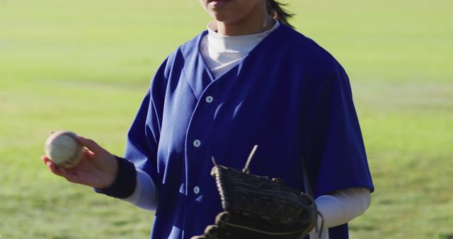 Female baseball player standing on a green field, dressed in blue uniform, holding a baseball in one hand and a glove in the other. Suitable for use in sports promotions, fitness campaigns, female athlete features, or outdoor activity advertisements.