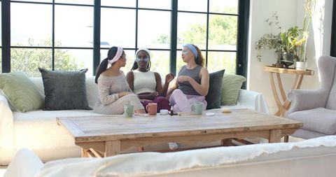Diverse women enjoying at-home spa day applying face masks, chatting in sunlit living room