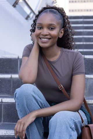 Smiling african woman sitting on urban steps wearing tee, jeans and brown crossbody bag
