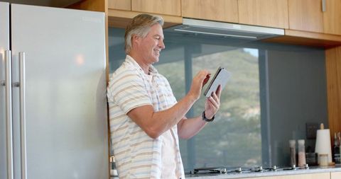 Mature man leaning on fridge tapping tablet in modern bright kitchen