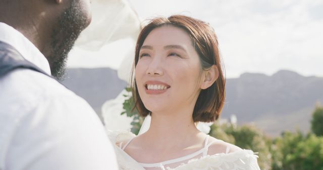 Image of happy diverse bride and groom smiling at each other at outdoor wedding. Marriage, love, happiness and inclusivity concept.