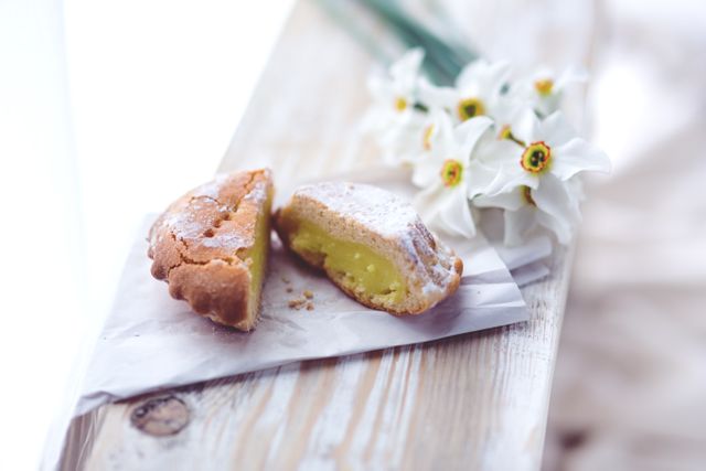 Elegant presentation of a delicious lemon pastry dusted with powdered sugar, placed on a piece of parchment paper alongside fresh spring daffodils on a wooden surface. Perfect for usage in culinary blogs, spring festival promotions, bakery shop advertisements, and social media sharing to convey freshness and delicacy.