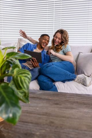 Laughing couple sharing tablet on cozy sofa, denim outfits, indoor greenery and natural light