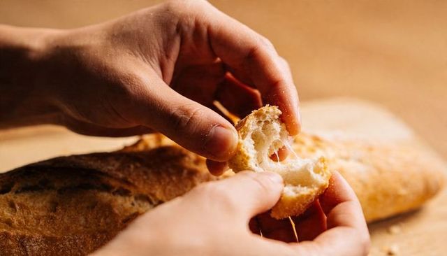 Hands tearing crusty baguette on wooden board, pulling apart airy artisan crumb