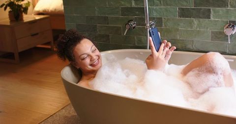 Relaxing african american woman soaking in bubble bath while smiling and using smartphone