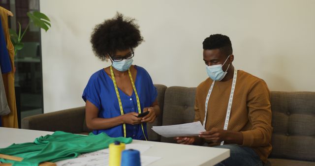 African american male and female fashion designers wearing masks taking photo of document. independent creative design business.