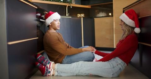 Couple sitting on kitchen floor holding hands wearing santa hats sharing holiday moment