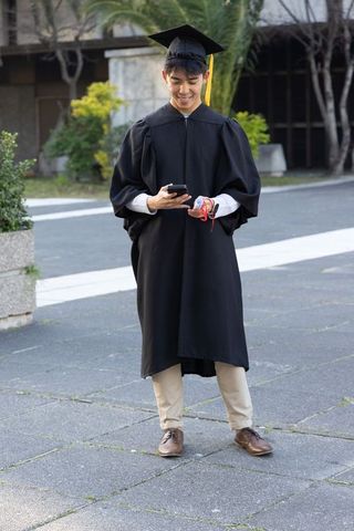 Asian graduate checking smartphone while holding red diploma cord in cap and gown on campus