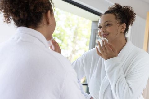 Smiling african american woman applying skincare with cotton pad at vanity window wearing white robe