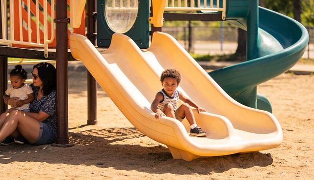 Curly toddler sliding down double yellow slide at sunny playground with caregiver nearby