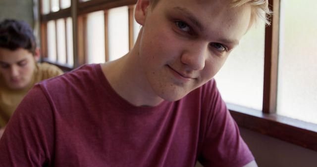 Picture of a teenage boy smiling while sitting in a cafe next to a window. Sunlight illuminates his face, creating a natural and relaxed atmosphere. He is wearing a red shirt. This image can be used to depict youth, happiness, relaxation, or a casual indoor setting for various marketing or inspirational projects.