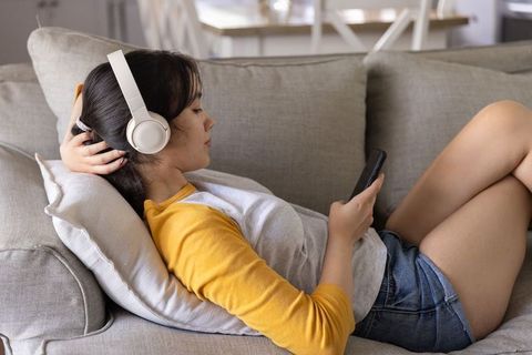 Asian woman reclining on sofa listening to headphones and using smartphone for leisure
