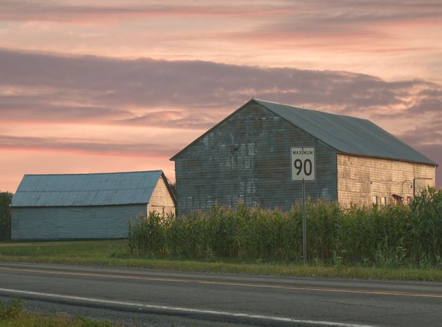 Scenic sunset view over rustic barns and road with speed limit sign in a rural area, providing a serene countryside ambiance. Useful for themes related to rural lifestyle, transportation speed regulations, farm life, and peaceful evening landscapes.