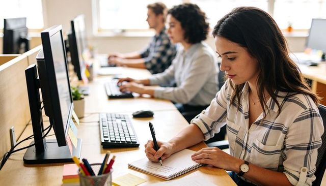 Young woman writing notes in open-plan office with coworkers at monitors, focused teamwork