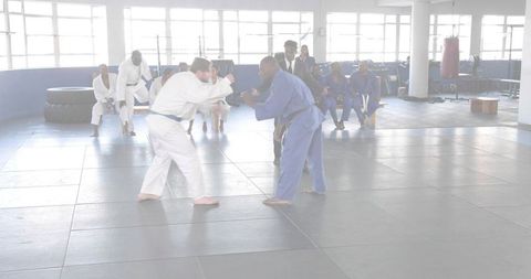 Grappling judo athletes sparring on dojo mat with referee and seated teammates