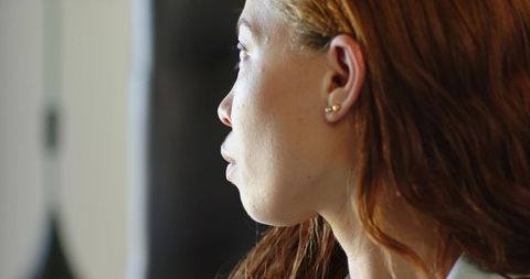 Pensive woman gazing out window closeup profile with auburn hair and gold stud earrings