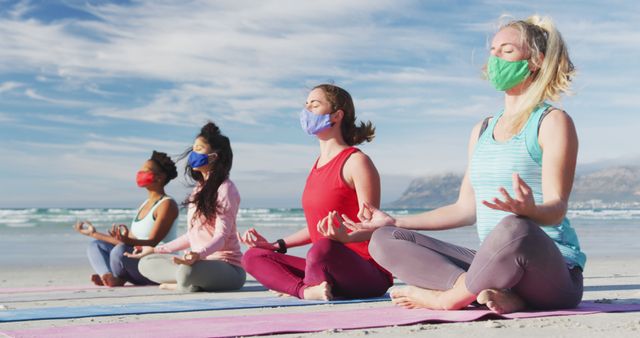 Group of diverse female friends wearing face masks meditating at the beach. healthy active lifestyle, outdoor fitness and wellbeing.