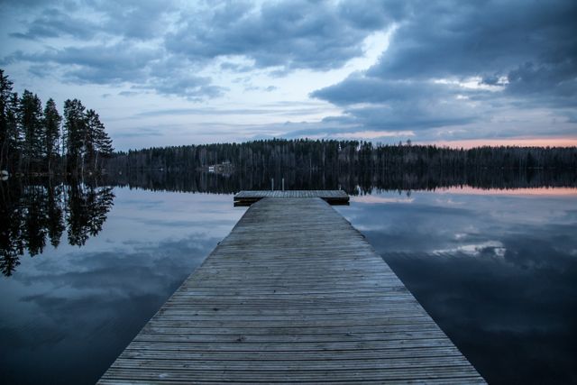 This picturesque scene showcases a wooden dock extending into a calm, reflective lake surrounded by trees at dusk. Ideal for use in travel brochures, nature conservation materials, or serene landscape collections, highlighting the tranquility and beauty of natural settings.