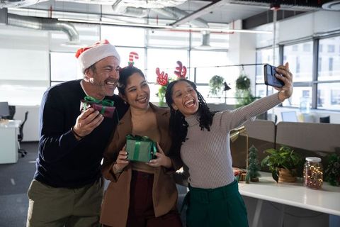 Coworkers celebrating office holiday party taking selfie with gifts and festive accessories