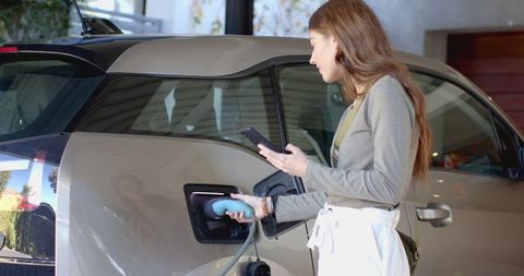 Woman Charging Electric Vehicle on Driveway Using Smartphone