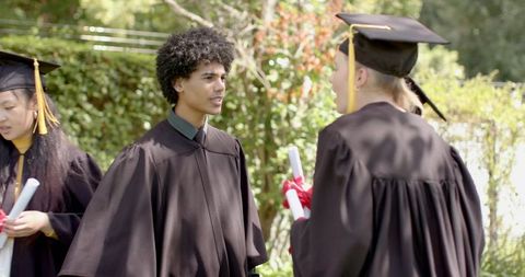 Diverse Graduates Wearing Black Gowns and Mortarboards Chatting on Campus Lawn