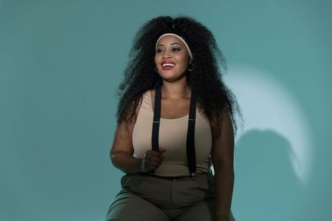 Smiling african american woman sitting in teal studio wearing headband and suspenders radiating conf