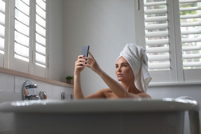 Woman sitting in a bathtub taking a selfie with a smartphone. She has a towel wrapped around her head. White shutters and modern bathroom fixtures are visible. Ideal for concepts of self-care, relaxation, modern lifestyle, and beauty routines.