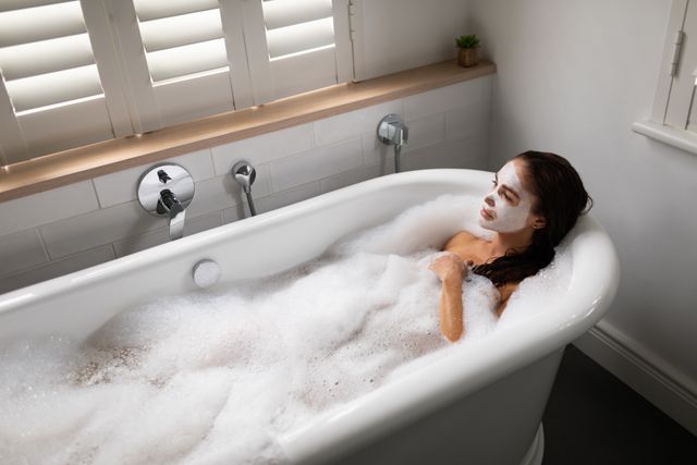 High-angle view of a beautiful woman enjoying a soothing bubble bath in a modern bathroom. She is wearing a face mask and appears calm and serene. This image is perfect for illustrating concepts of self-care, relaxation, spa treatments, and personal wellness routines. Ideal for use in beauty and wellness blogs, spa advertisements, and health and lifestyle magazines.