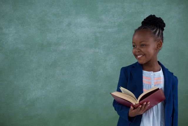 Young girl standing in classroom holding book and smiling. Ideal for educational content, school promotions, academic materials, and childhood learning resources.