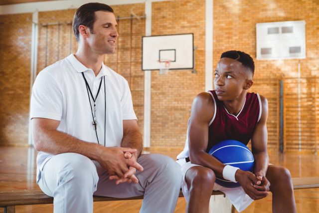 Smiling coach guiding basketball player while sitting on bench in court