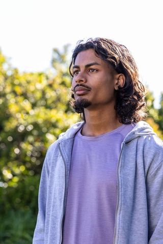 Young man standing in sunlit park wearing gray hoodie and lavender t-shirt gazing upward