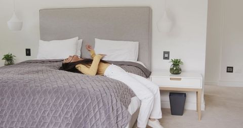 Woman Lying Back on Bed in Minimalist Bedroom Wearing Mustard Top and White Pants