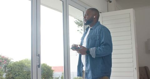 Man listening to wireless earbuds while checking smartphone by sunlit sliding glass doors