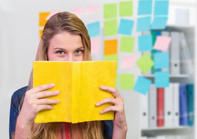 Young woman holding a yellow book, partially covering her face, with colorful sticky notes on the wall in the background. Ideal for use in educational, organizational, and creative planning contexts. Perfect for illustrating concepts of studying, focus, and office work.
